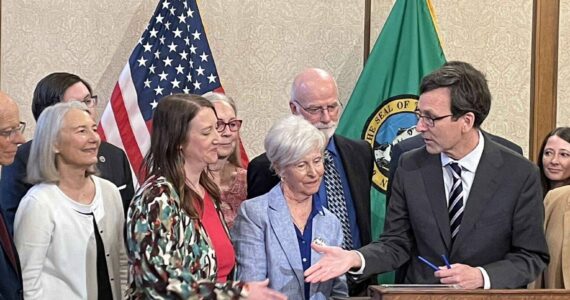 Gov. Bob Ferguson, at podium, goes to shake hands with state Sen. Noel Frame, D-Seattle, at the signing of a bill to make clergy mandatory reporters of child abuse and neglect, on May 2, 2025 in Olympia. At center is Mary Dispenza, a founding member of the Catholic Accountability Project. (Photo by Jerry Cornfield/Washington State Standard)
