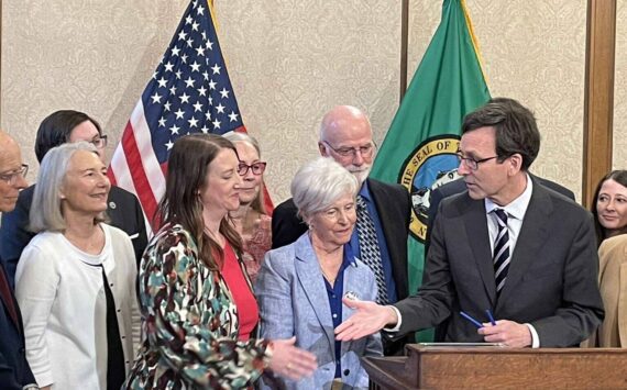 Gov. Bob Ferguson, at podium, goes to shake hands with state Sen. Noel Frame, D-Seattle, at the signing of a bill to make clergy mandatory reporters of child abuse and neglect, on May 2, 2025 in Olympia. At center is Mary Dispenza, a founding member of the Catholic Accountability Project. (Photo by Jerry Cornfield/Washington State Standard)
