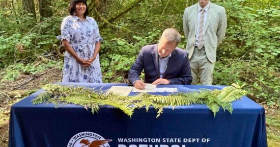 Washington Public Lands Commissioner Dave Upthegrove signs a directive ordering the conservation of about 77,000 acres of older forests on state logging lands, on Aug. 26, 2025, at the High Point Trailhead on Tiger Mountain. Behind him are Washington Conservation Action CEO Alyssa Macy, left, and Thurston County Commissioner Tye Menser, right. (Photo by Emily Fitzgerald/Washington State Standard)
