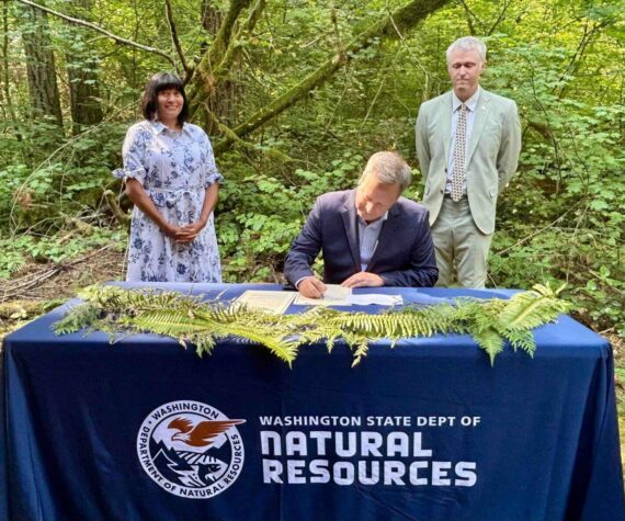 Washington Public Lands Commissioner Dave Upthegrove signs a directive ordering the conservation of about 77,000 acres of older forests on state logging lands, on Aug. 26, 2025, at the High Point Trailhead on Tiger Mountain. Behind him are Washington Conservation Action CEO Alyssa Macy, left, and Thurston County Commissioner Tye Menser, right. (Photo by Emily Fitzgerald/Washington State Standard)