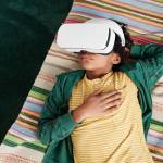 High angle view of African little boy relaxing on sofa and using VR goggles to watch virtual reality video during his leisure time at home