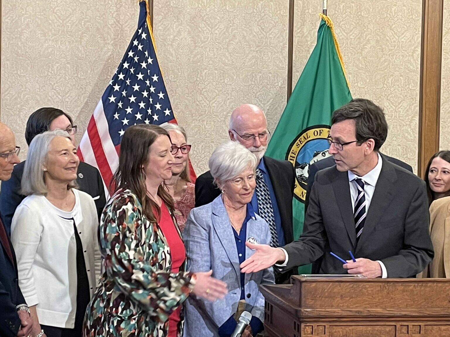 Gov. Bob Ferguson, at podium, goes to shake hands with state Sen. Noel Frame, D-Seattle, at the signing of a bill to make clergy mandatory reporters of child abuse and neglect, on May 2, 2025 in Olympia. At center is Mary Dispenza, a founding member of the Catholic Accountability Project. (Photo by Jerry Cornfield/Washington State Standard)