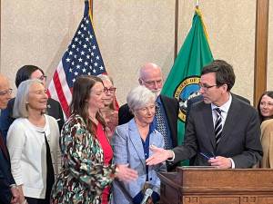 Gov. Bob Ferguson, at podium, goes to shake hands with state Sen. Noel Frame, D-Seattle, at the signing of a bill to make clergy mandatory reporters of child abuse and neglect, on May 2, 2025 in Olympia. At center is Mary Dispenza, a founding member of the Catholic Accountability Project. (Photo by Jerry Cornfield/Washington State Standard)